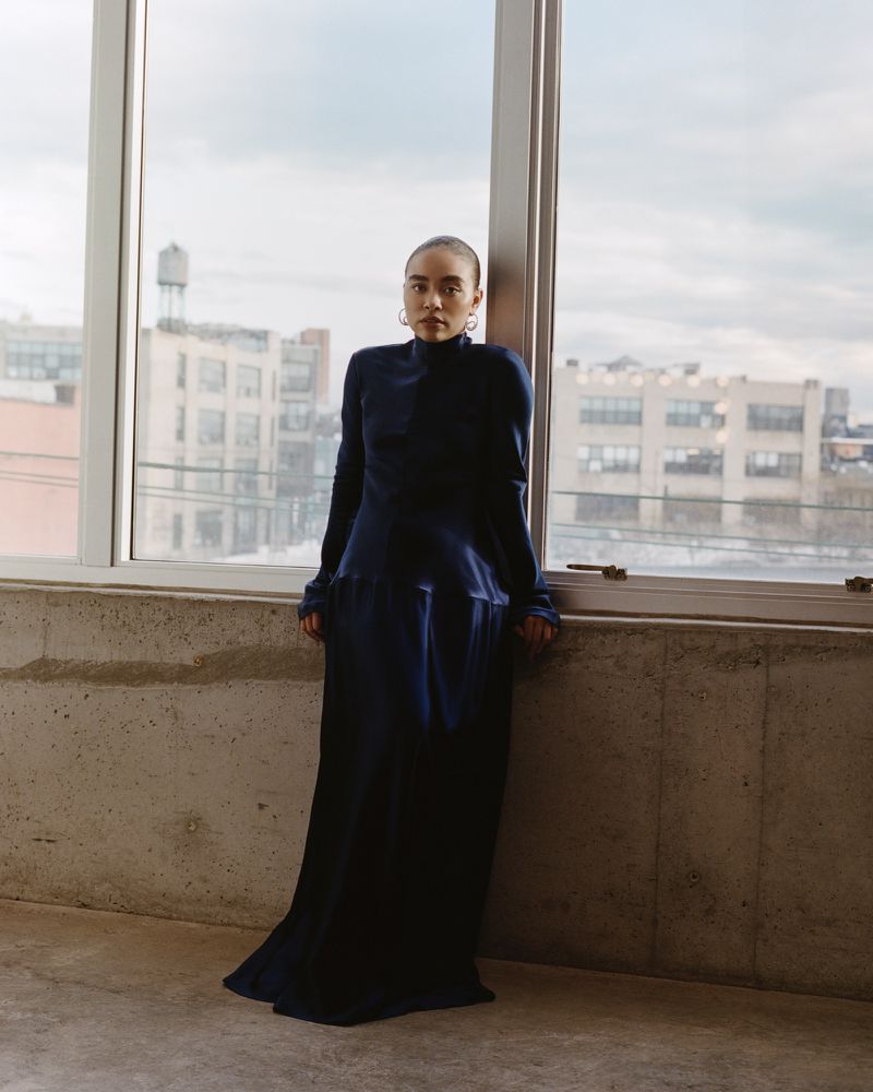 Woman in long blue dress standing by window in urban loft with city views, minimalist style, natural light portrait.
