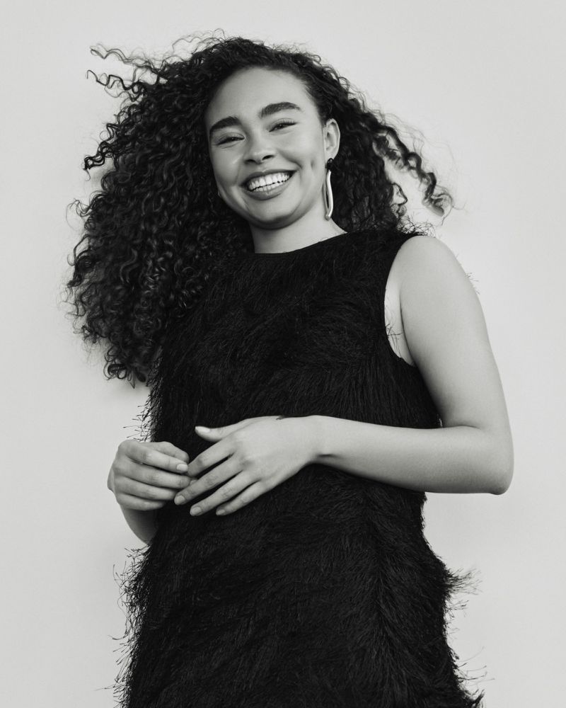 Portrait of a Black woman with curly hair wearing a sleeveless black dress in a studio setting, smiling confidently at the camera.