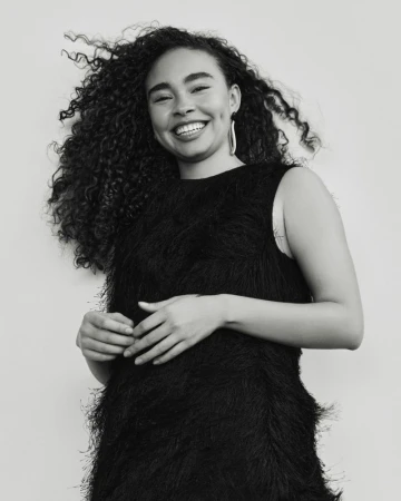 Portrait of a Black woman with curly hair wearing a sleeveless black dress in a studio setting, smiling confidently at the camera.