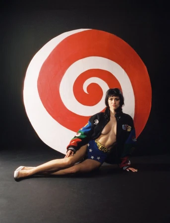 A woman in a colorful jacket and star-patterned shorts poses in front of a large, red and white spiral target against a black backdrop.
