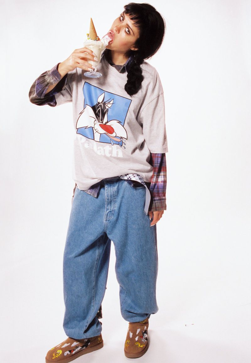 A young adult female stands in a white studio, eating ice cream from a tall glass. She is wearing casual clothing with a graphic tee.