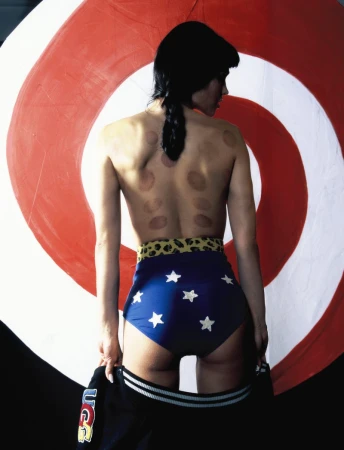 Adult female model poses against a bullseye in swim bottoms with stars, facing away from the camera, with a jacket in hand.