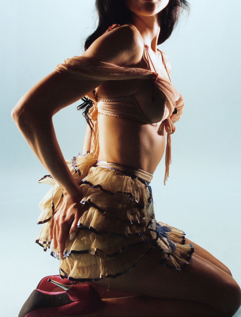 Woman in lingerie kneeling indoors with red high heels in a studio with a light blue background, arms raised above her shoulders.