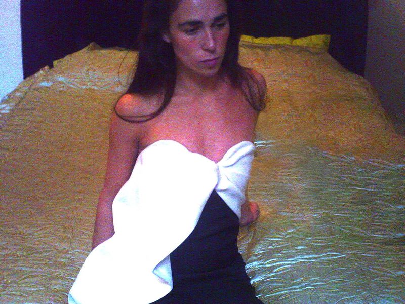 Adult female in a black dress with a large white bow poses indoors on a gold-colored bed with a dark purple wall behind it.