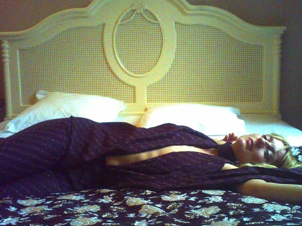 Woman lying on a floral patterned bed with white pillows and a light colored headboard with an oval design in a neutral toned room.