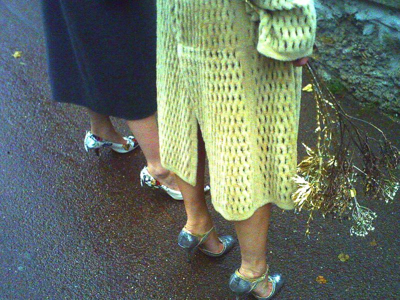 Two women stand on wet pavement, one in a yellow knitted coat and silver heels, the other in a dark skirt and floral print heels.