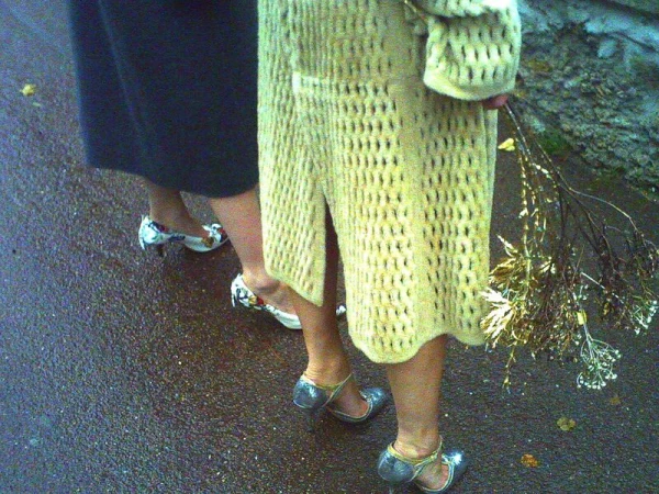 Two women stand on wet pavement, one in a yellow knitted coat and silver heels, the other in a dark skirt and floral print heels.