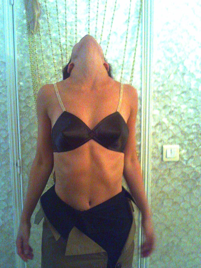 Adult female posing with head tilted back, wearing bra and clothing around waist. Background features gold chain curtain and tiled wall.