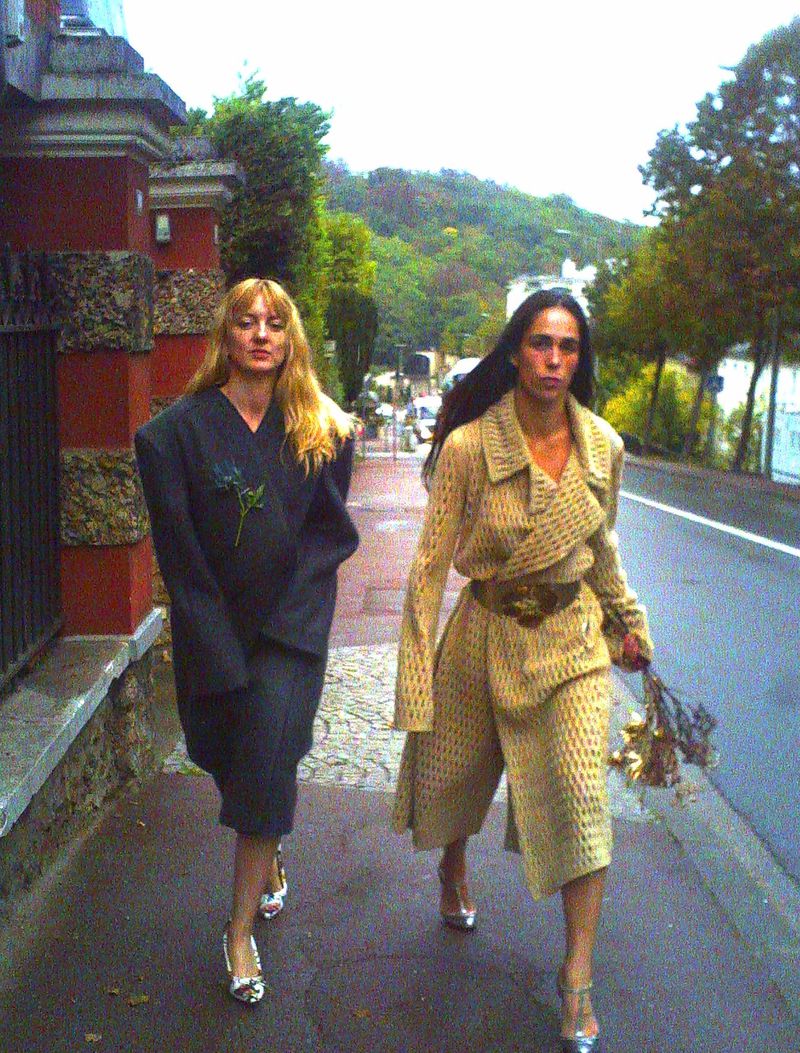 Two women walking on a city street, both wearing distinctive coats and shiny heels, with trees and buildings in the background.