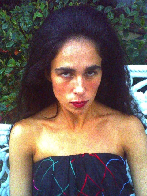 Portrait of a woman with long dark hair and red lipstick wearing a colorful strapless dress, posing outdoors against a green leafy background.