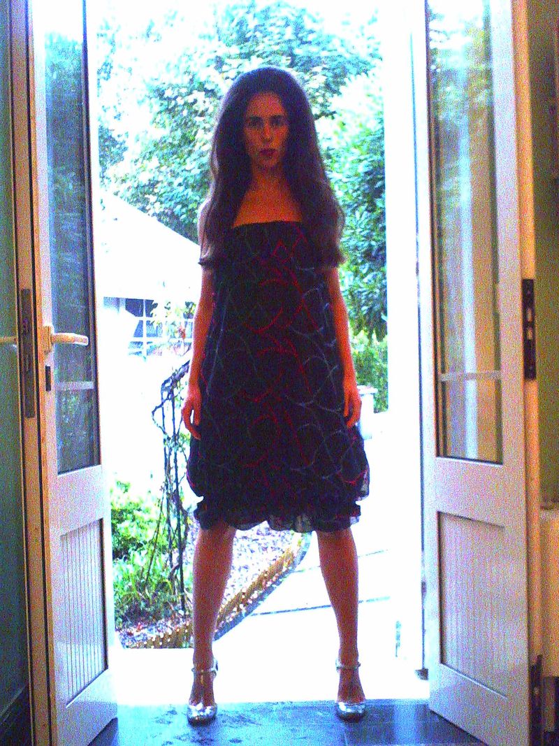 A woman stands in a doorway wearing a strapless dress and heels, with a yard visible through the open door. The focus is on her fashion.