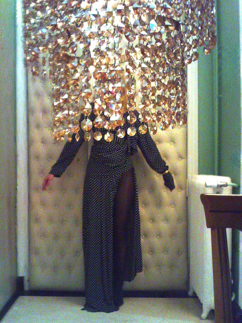 Person in long polka-dot dress stands behind a gold crystal beaded curtain in a dimly lit interior space with chair in background.