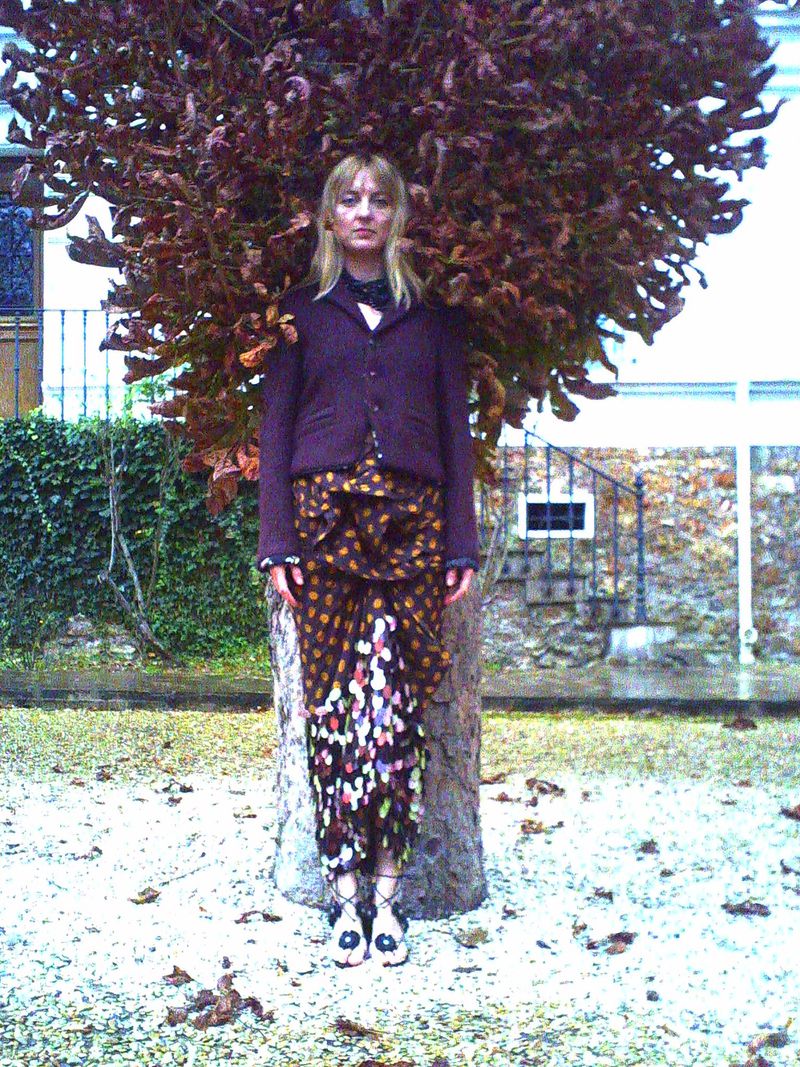 Adult female in outdoor fashion portrait. Woman in jacket and patterned skirt stands near a tree against a stone building backdrop.