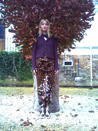 Adult female in outdoor fashion portrait. Woman in jacket and patterned skirt stands near a tree against a stone building backdrop.