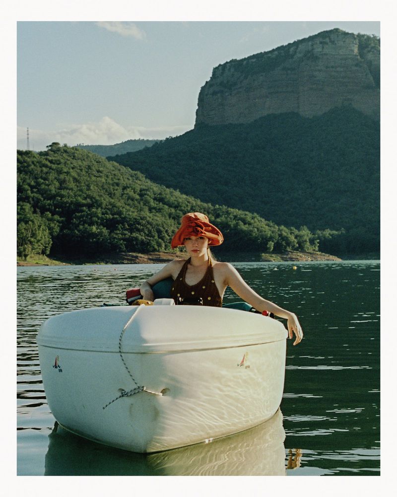 A woman in a boat on a lake, wearing a hat and swimsuit, with a large rock formation in the background on a sunny day.