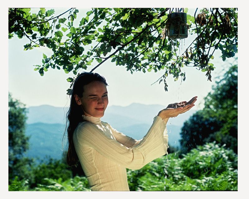 Young adult female winks, holding hands under water droplets falling from a basket, set against a backdrop of mountains and green foliage.