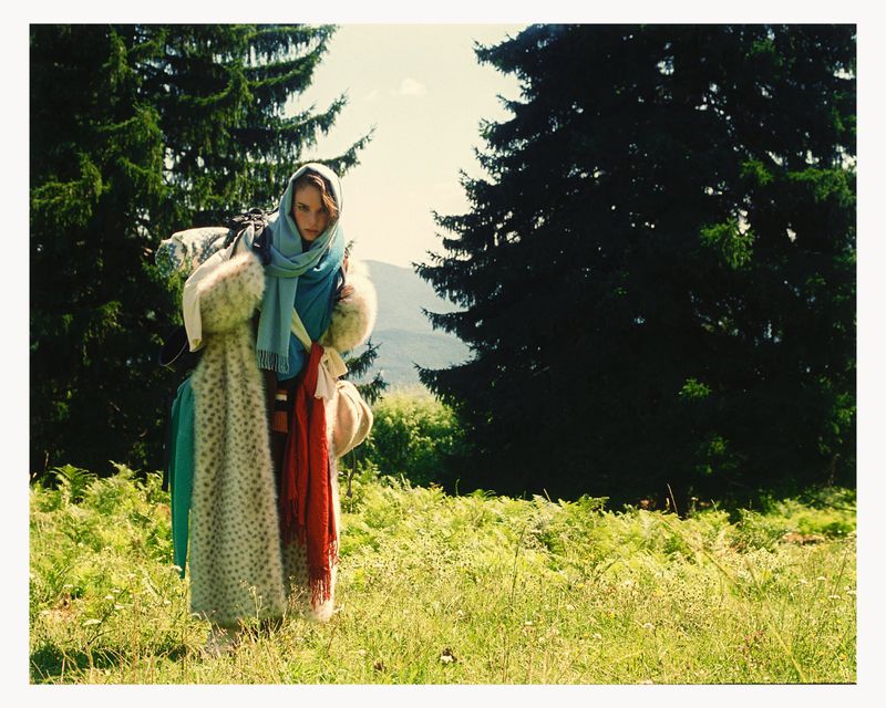 Woman in headscarf stands in field near forest wearing fur and fabric wraps, illuminated by sunlight, fashion portrait, outdoor setting.