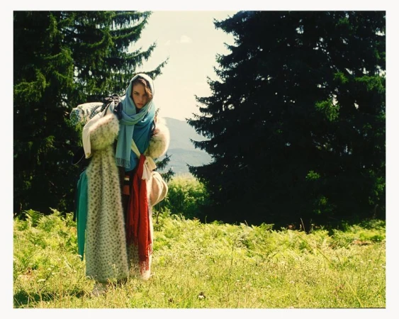 Woman in headscarf stands in field near forest wearing fur and fabric wraps, illuminated by sunlight, fashion portrait, outdoor setting.