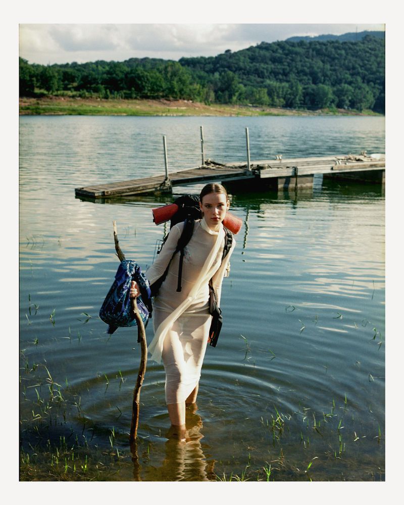 A woman in a light dress wading through a lake with a backpack and walking stick, with forested hills and a cloudy sky in the distance.