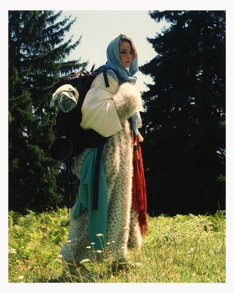 Woman in patterned coat, headscarf, and backpack standing in a field with trees, shot in natural light with a documentary style.