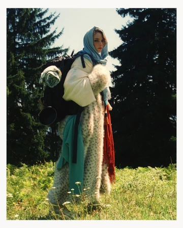 Woman in patterned coat, headscarf, and backpack standing in a field with trees, shot in natural light with a documentary style.