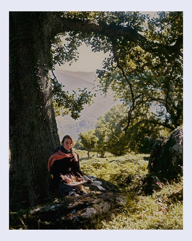 Woman sitting against large tree trunk in green forest, sketching in notebook under sunlight with mountain range in background.
