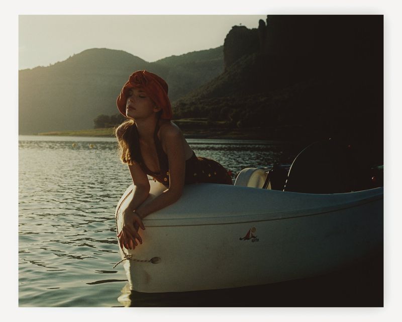 Woman in hat poses in canoe on lake with mountains in background in outdoor fashion shot taken on sunny day with soft lighting.