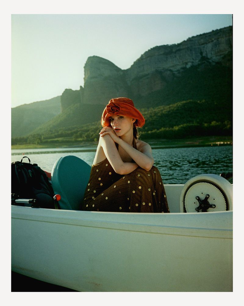 Woman in brown polka-dot dress and orange hat sits in a boat on a lake with mountains in the background on a sunny day.