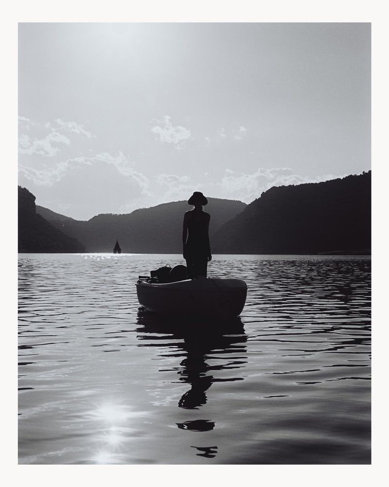 Black and white photo of a woman in a hat standing in a small boat on a calm lake with silhouetted mountains in the background.