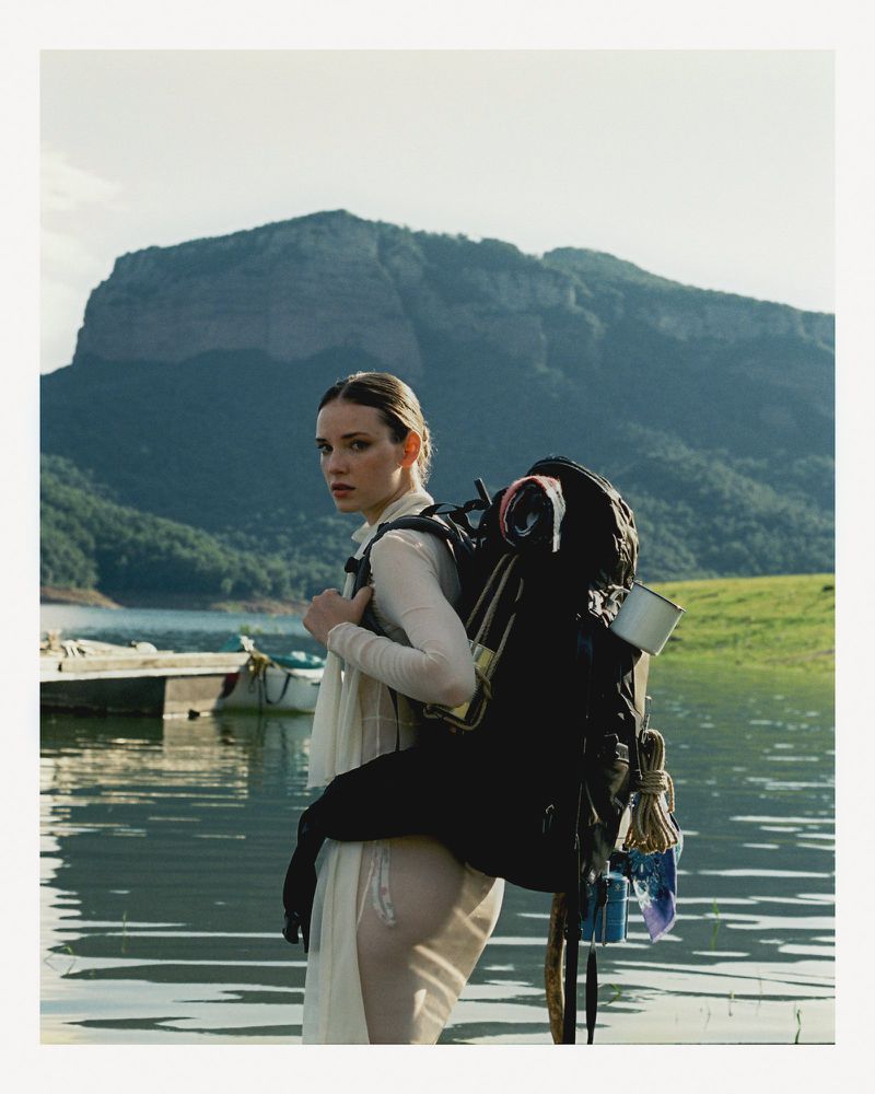 Woman in a light dress carrying a large hiking backpack wading in water with a mountain range and lake visible in the background.