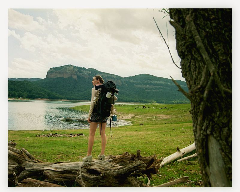 Young woman with backpack standing on a fallen tree trunk, looking out at a lake and mountains in the background on an overcast day.