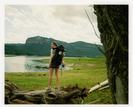 Young woman with backpack standing on a fallen tree trunk, looking out at a lake and mountains in the background on an overcast day.