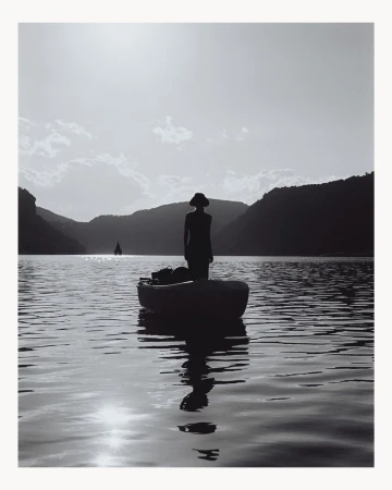 Silhouette of a person standing in a small boat on a calm lake surrounded by hills under a bright sky, photographed by Deid Garcia.