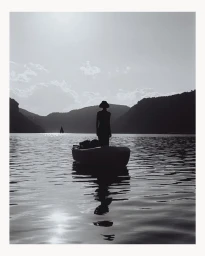 Silhouette of a person standing in a small boat on a calm lake surrounded by hills under a bright sky, photographed by Deid Garcia.