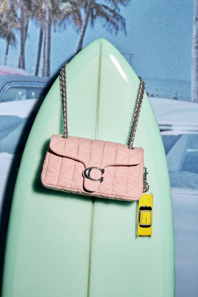 Pale pink Coach bag with silver chain strap on a light green surfboard, beach scene backdrop with palm trees and a vintage car.