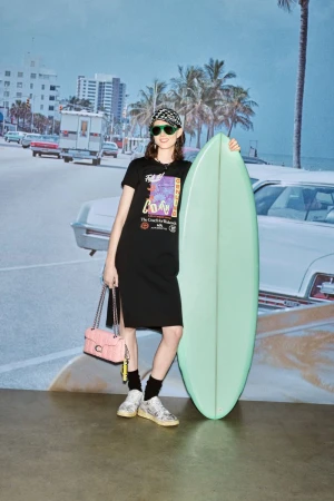 Adult female in casual dress posing with a surfboard in front of a tropical mural backdrop. She wears sunglasses and a patterned headscarf.