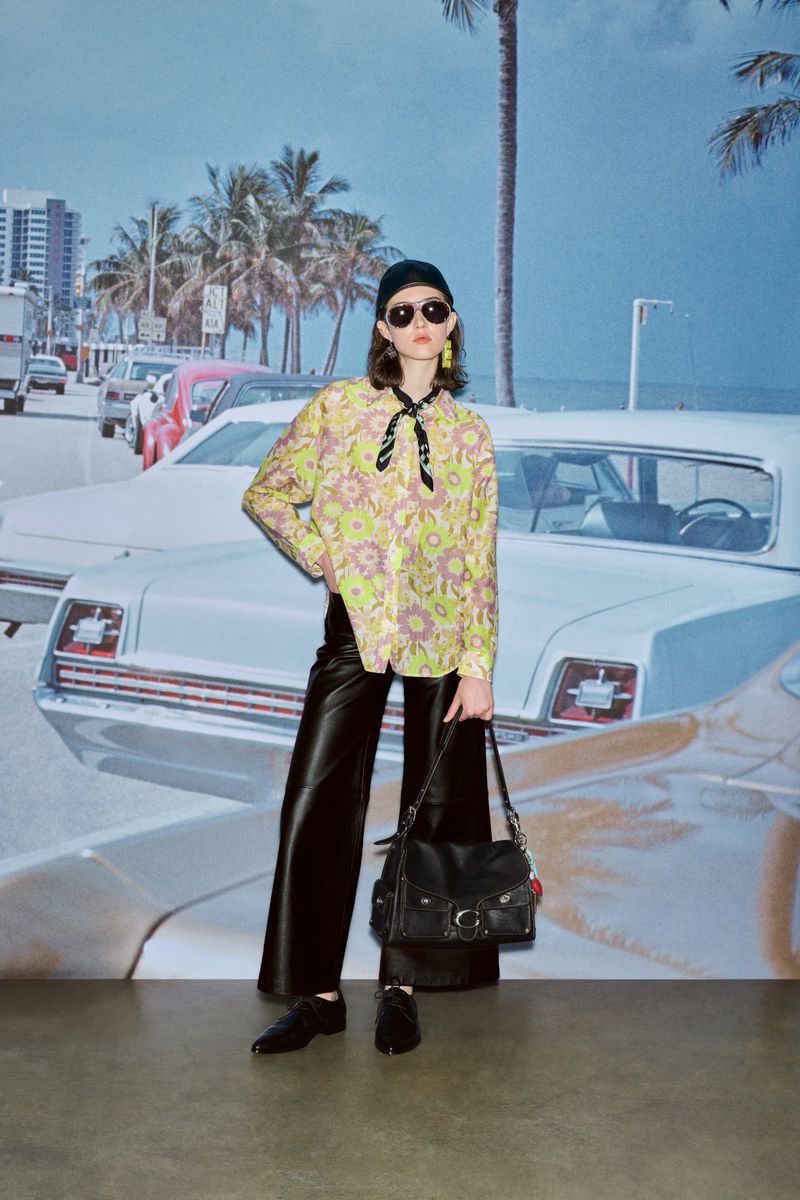 A woman in fashionable clothing poses in front of a backdrop featuring a sunny beach scene with palm trees and vintage cars.