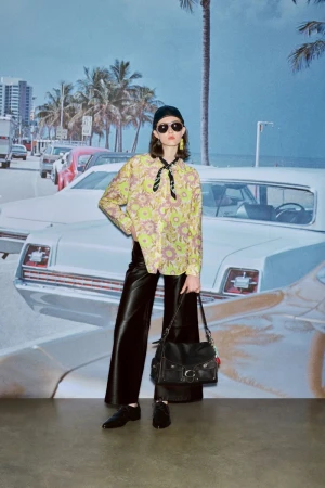 A woman in fashionable clothing poses in front of a backdrop featuring a sunny beach scene with palm trees and vintage cars.