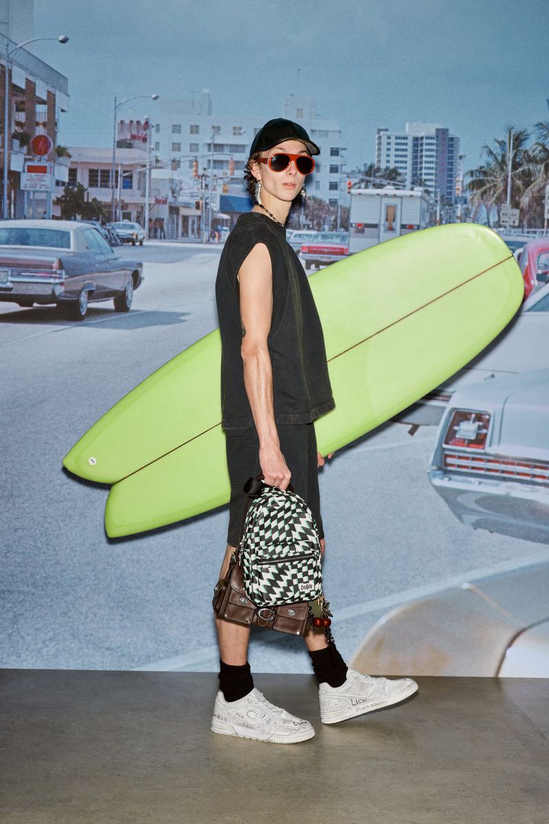 Stylish male model poses with surfboard and backpacks against a vintage city backdrop. Fashion, style, and summer vibes.