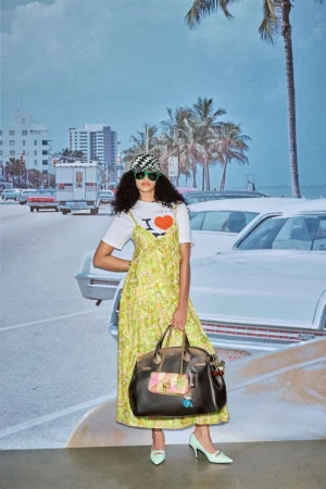 An adult female model wearing a patterned dress, accessories, and shoes, holding a handbag in front of a beach backdrop with cars and palm trees.