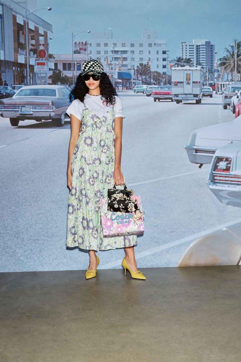 A woman poses with handbags on a street backdrop, wearing a floral dress, white shirt, and yellow heels, accessorized with sunglasses and a cap.