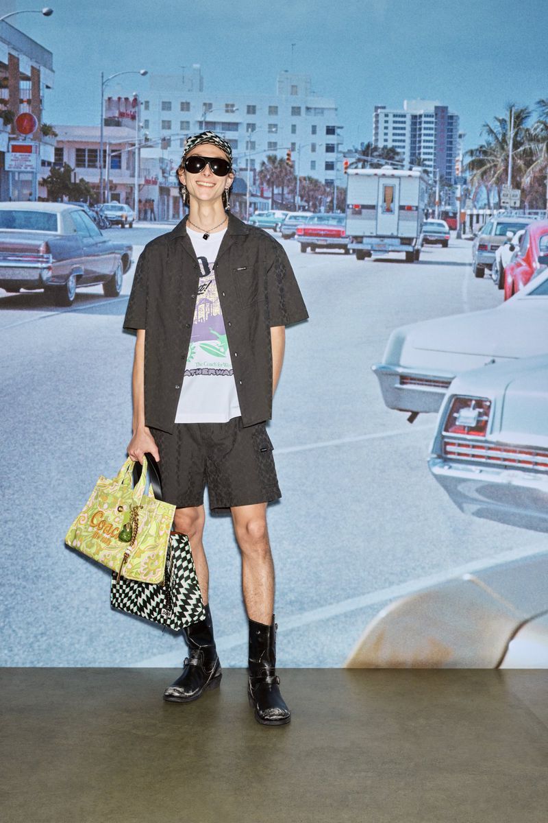 A fashionable young man in black shorts and boots stands in front of a Miami street scene holding two bags with neutral expression.