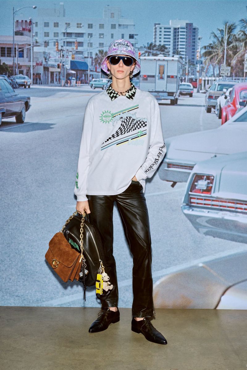 Fashionable adult posing in a long-sleeve graphic shirt, black pants, bucker hat, sunglasses, and backpack against a backdrop with city street.