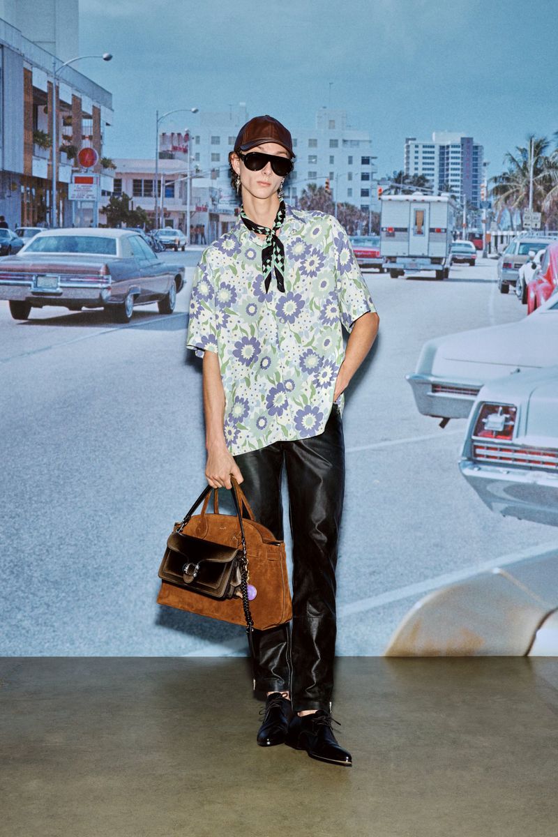 Adult male fashion model wears a floral shirt, dark pants, sunglasses, and hat, holding a purse in an outdoor street setting.