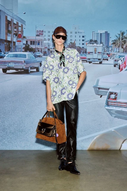 Adult male fashion model wears a floral shirt, dark pants, sunglasses, and hat, holding a purse in an outdoor street setting.