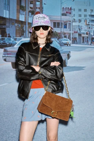 Woman poses with brown Coach purse, wearing a Coach bucket hat, leather jacket, and printed shorts, with a city backdrop.