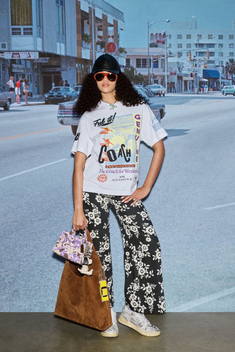 A woman in a Coach t-shirt, floral pants, bucket hat, and orange sunglasses is posing with Coach bags in front of a backdrop.
