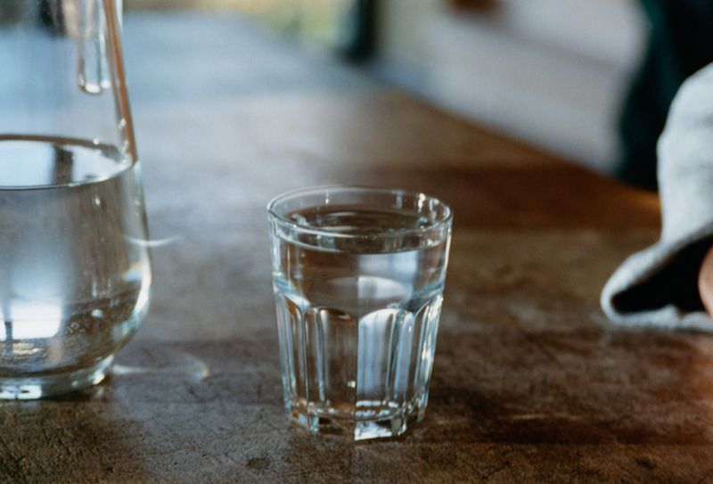 Still life of a clear drinking glass filled with water next to a glass carafe on a wooden surface indoors, with soft lighting.