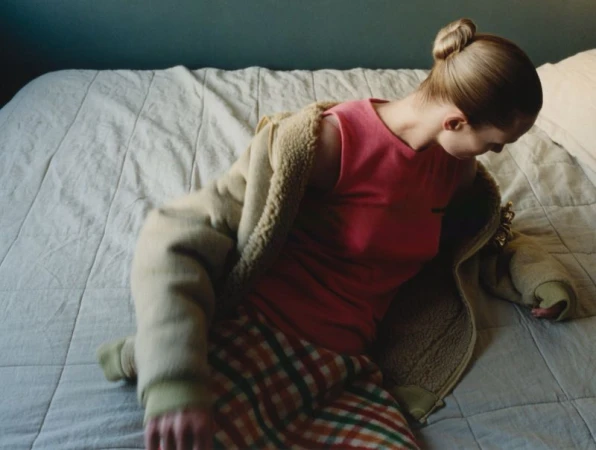 Young woman with blonde hair in a bun, sitting indoors on a bed with white linens wearing a pink top, jacket, and plaid skirt.