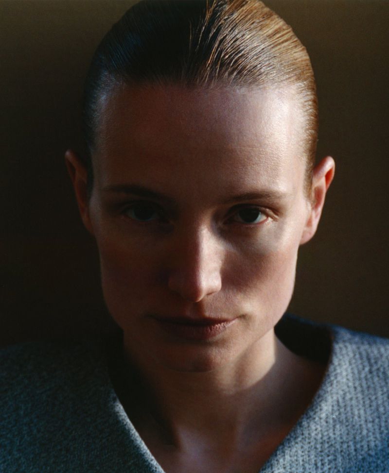 Close-up portrait of an adult woman with slicked-back short hair, wearing a grey top, set against a dark, gradient background.